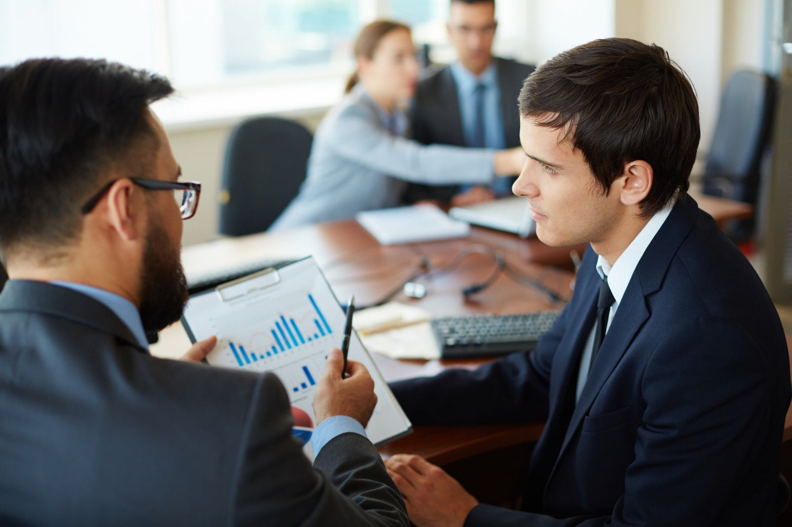 A close-up of a commercial finance operator in a dark suit listening to a colleague holding a clipboard with bar graphs, reviewing cost and performance metrics for inbound live transfer campaigns.