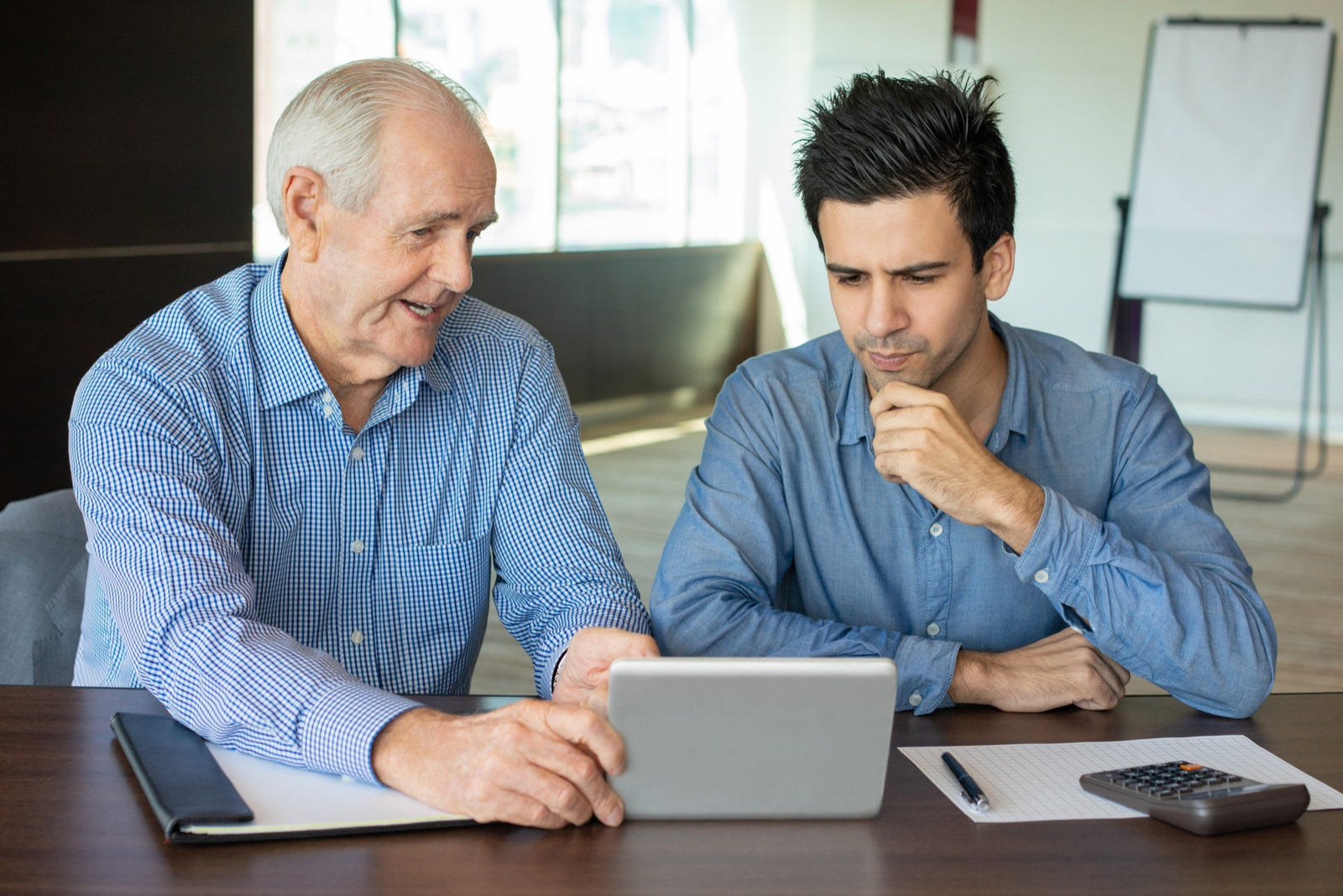A senior funding broker demonstrating business loan options on a digital tablet to a pensive business owner looking to secure working capital.