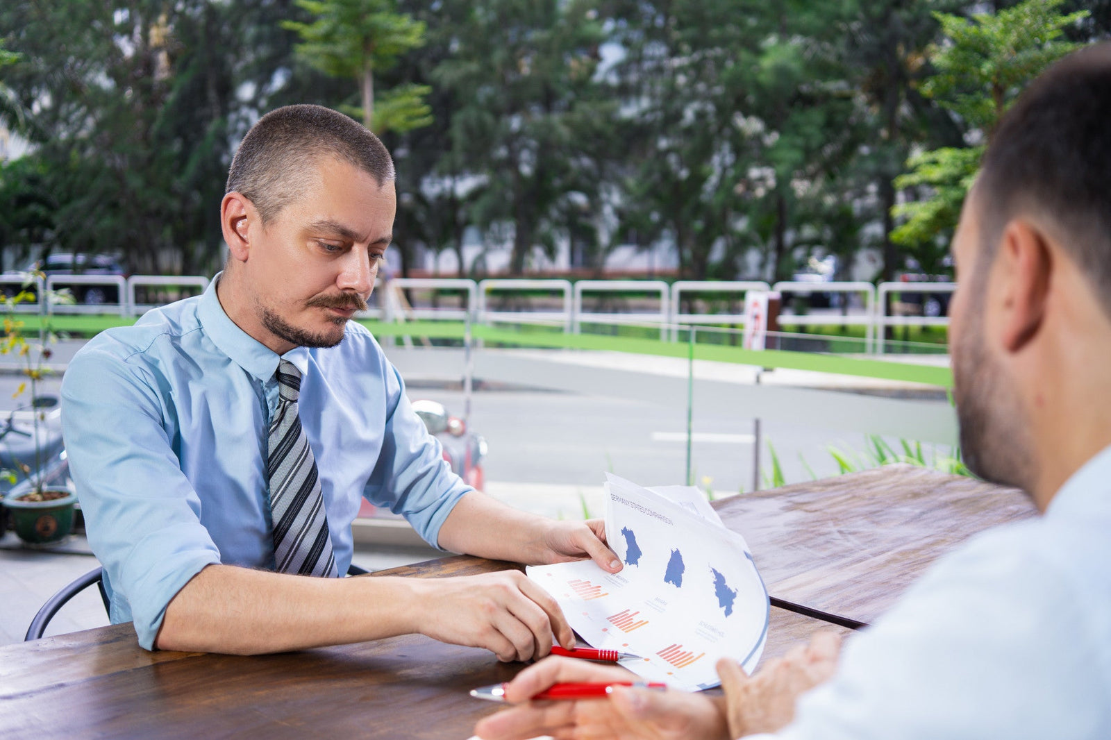 A commercial finance manager holding a red pen and reviewing a financial chart and price breakdown document at an outdoor office table during a client consultation.