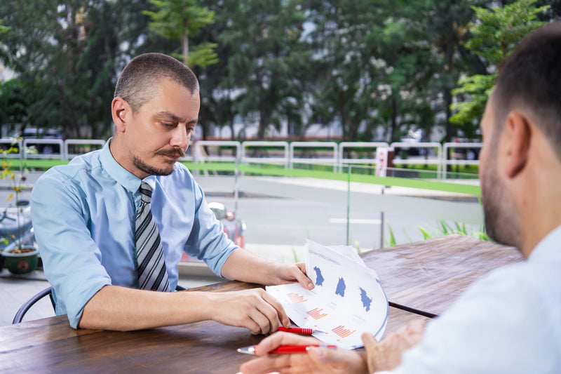 A commercial finance manager holding a red pen and reviewing a financial chart and price breakdown document at an outdoor office table during a client consultation.
