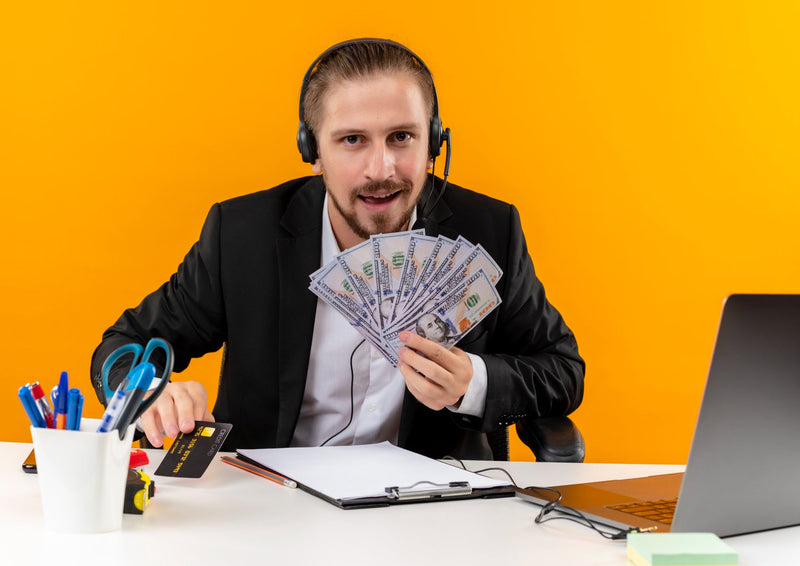 A merchant cash advance broker wearing a headset holding cash and a credit card at his desk, representing successful live transfer business loan leads