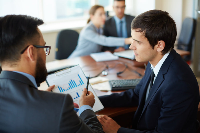 A close-up of a commercial finance operator in a dark suit listening to a colleague holding a clipboard with bar graphs, reviewing cost and performance metrics for inbound live transfer campaigns.