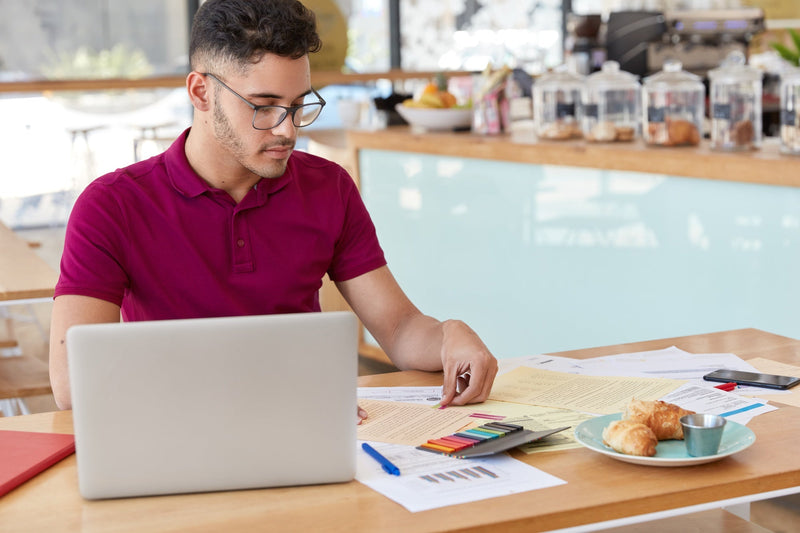 A business operator wearing glasses and a magenta polo shirt sits at a wooden table reviewing printed financial data documents, charts, and public record filings next to an open laptop.
